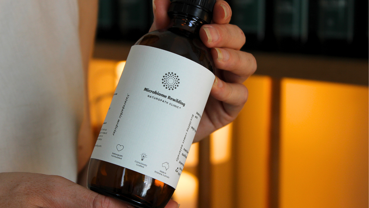 Microbiome Rewilding Naturopath Clinic Brisbane Australia Online Appointments experienced Naturopath holding a bottle of a custom Microbiome Rewilding Naturopath Clinic Herbal Medicine Tonic on a wooden table with other naturopathic dispensary and skincare items for personalised holistic women’s health