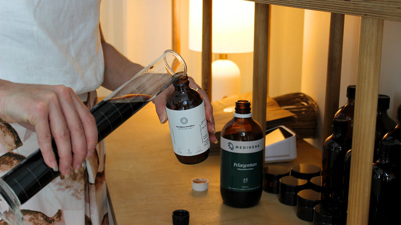 Microbiome Rewilding Naturopath Clinic Brisbane Australia Online Appointments experienced Naturopath holding a bottle of a custom Microbiome Rewilding Naturopath Clinic Herbal Medicine Tonic on a wooden table with other naturopathic dispensary and skincare items for personalised holistic women’s health