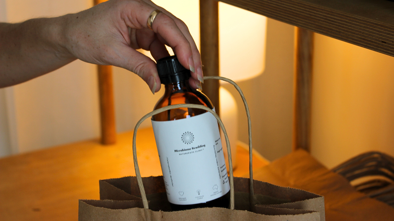 Microbiome Rewilding Naturopath Clinic Brisbane Australia Online Appointments experienced Naturopath holding a bottle of a custom Microbiome Rewilding Naturopath Clinic Herbal Medicine Tonic on a wooden table with other naturopathic dispensary and skincare items for personalised holistic women’s health